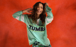 Female model posing playfully in front of a large textured orange backdrop in a ZUMBA polo shirt.